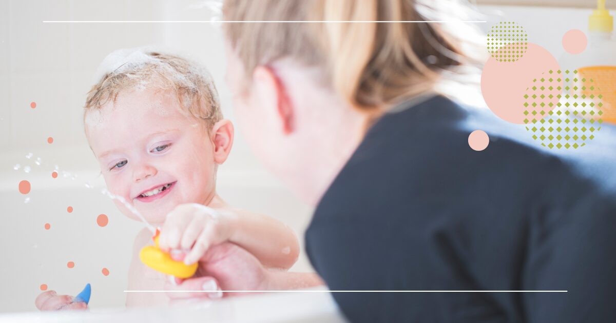Smiling toddler playing with a rubber duck in the bathtub while an adult watches nearby.